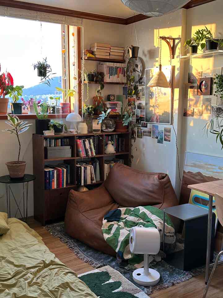 温かみのある木目調と豊富な緑の植物が調和した小さな部屋、ビーズクッションソファ、本棚、壁棚が配置された自然で居心地の良いインテリア