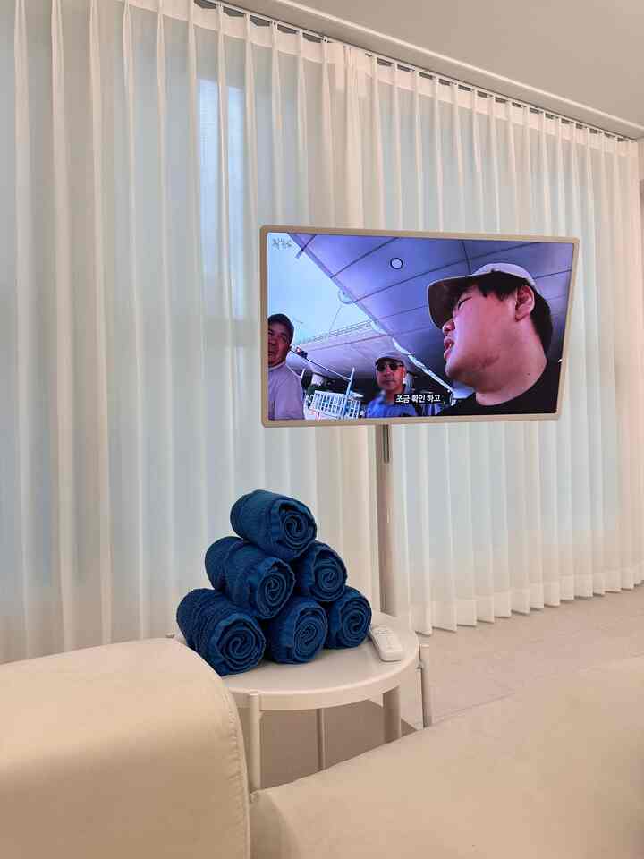 ホワイトとブルーのトーンのリビングルーム、シフォンカーテンとハンセムのソファ、イケアのナイトテーブルに丸めた青いタオルとスタンドバイミーのテレビが配置された清潔で落ち着いた雰囲気のインテリア