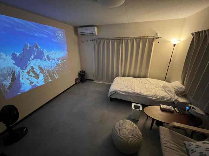 白とグレーのトーンの1DK寝室、ベッドとプロジェクタースクリーン、カーテン、アームチェアが配置されたシンプルで落ち着いた雰囲気の部屋