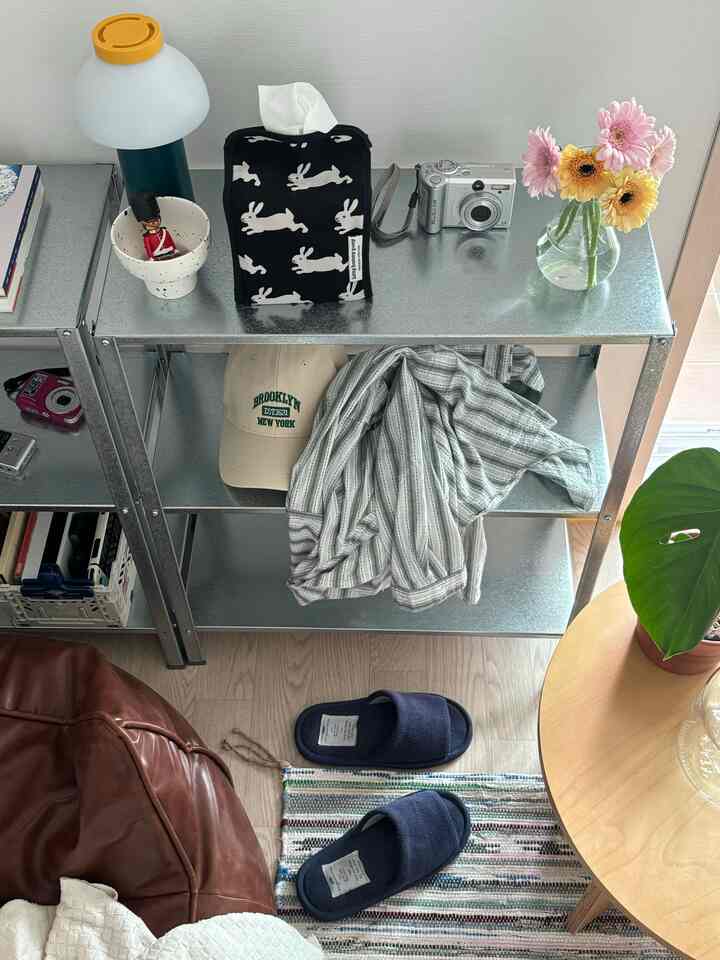 ナチュラルな色調のワンルーム、銀色のイケア棚とブラウンのビーズクッション、パステルカラーの花の入ったガラス花瓶が配置された印象的な空間