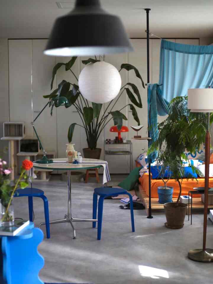 Blue and orange toned living-bedroom featuring mid-century modern round table and stools, abundant plants creating a cozy atmosphere