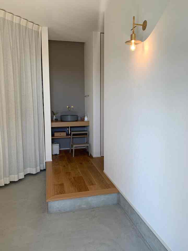 ナチュラルカラーの玄関空間、左にリネンカーテン、右に壁付照明、中央に木製フローリングと多目的洗面台が配置されたミニマルなインテリア