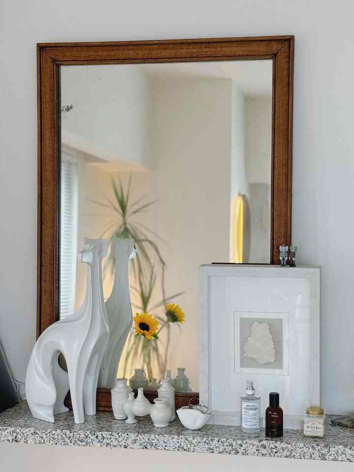 Natural-toned wood frame mirror with white vases and elegant objets displayed on a granite ledge in cozy living room corner