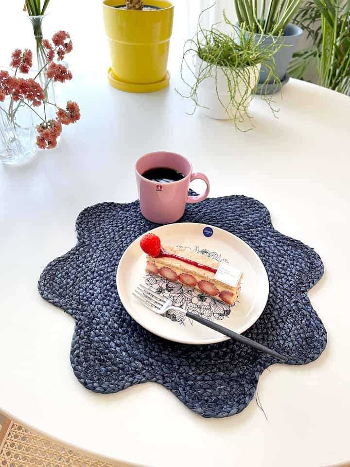 白いテーブルの上にピンクのマグカップとイチゴケーキが置かれた、黒い花形ランチョンマットのある雰囲気のあるおうちカフェ