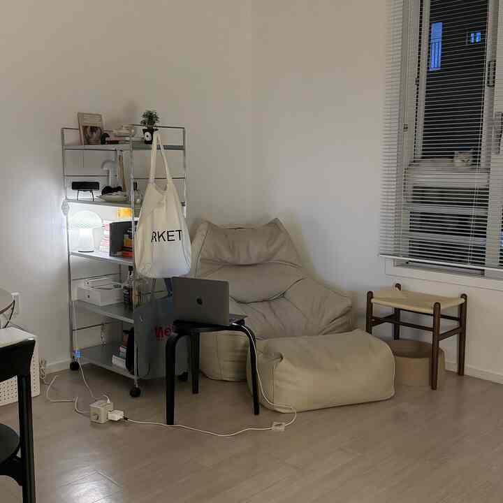 白い壁と木目調の床が特徴のミニマルなリビング、ベージュのビーズクッションと金属製の棚、窓辺に猫がいる落ち着いた雰囲気のインテリア