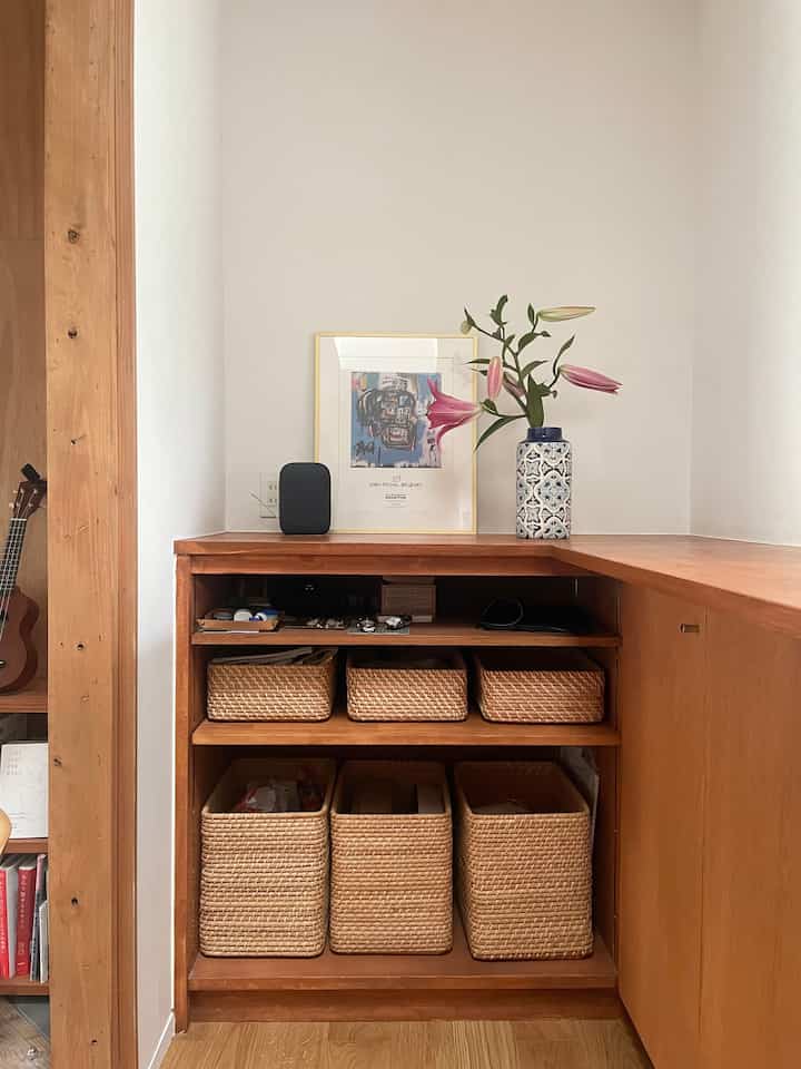 Natural wood-tone kitchen corner storage featuring Basquiat art, large lily flower vase, and tidy rattan baskets on white walls