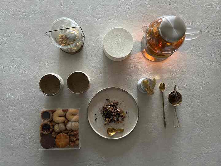 Charcoal-toned table featuring transparent glass teapot, teacups, and gold spoon arranged in a cozy home cafe setting