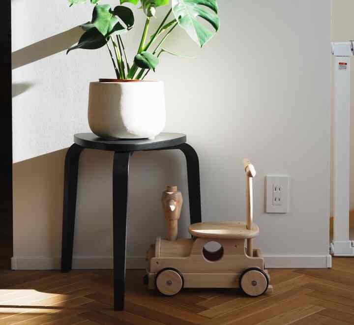 ナチュラルな木目調のヘリンボーン床に、モダンな黒いスツールの上に置かれた植物と木製の子供用乗り物おもちゃがある静かな子供部屋のインテリア