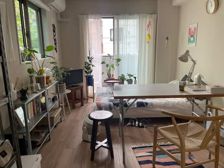 ナチュラルな木目調の床と白いカーテン、多数の植物とシンプルな家具が配置された1Kのワンルーム