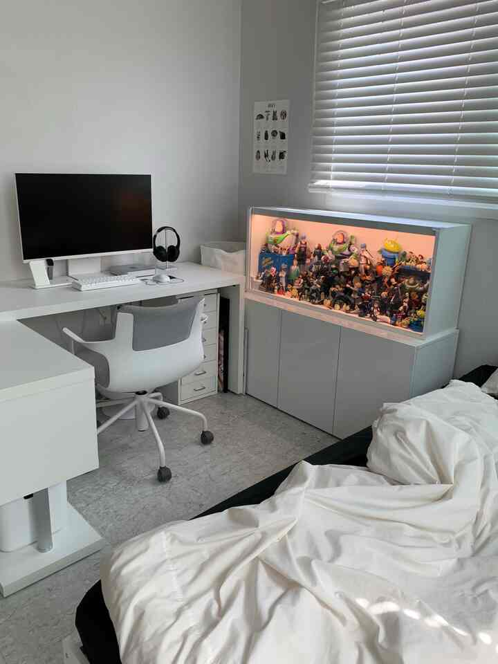 白とグレーのトーンでまとめられたコンパクトなホームオフィス、デスクとオフィスチェア、フィギュアが配置された照明付きの飾り棚があるすっきりとした空間