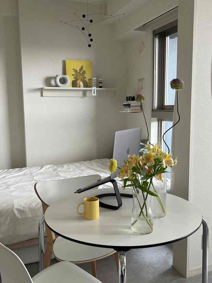 白い壁と家具が配されたシンプルな一人暮らしの空間、丸テーブル上に黄色の小物と花が暖かい雰囲気を演出