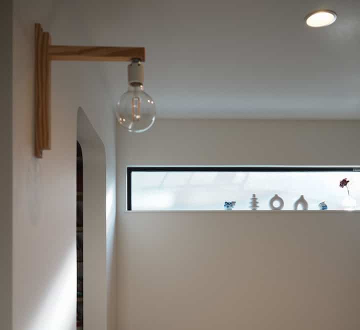 白い壁と木製ブラケットライトがあるシンプルな玄関、自然光が差し込む細長い高窓のある雰囲気のあるインテリア