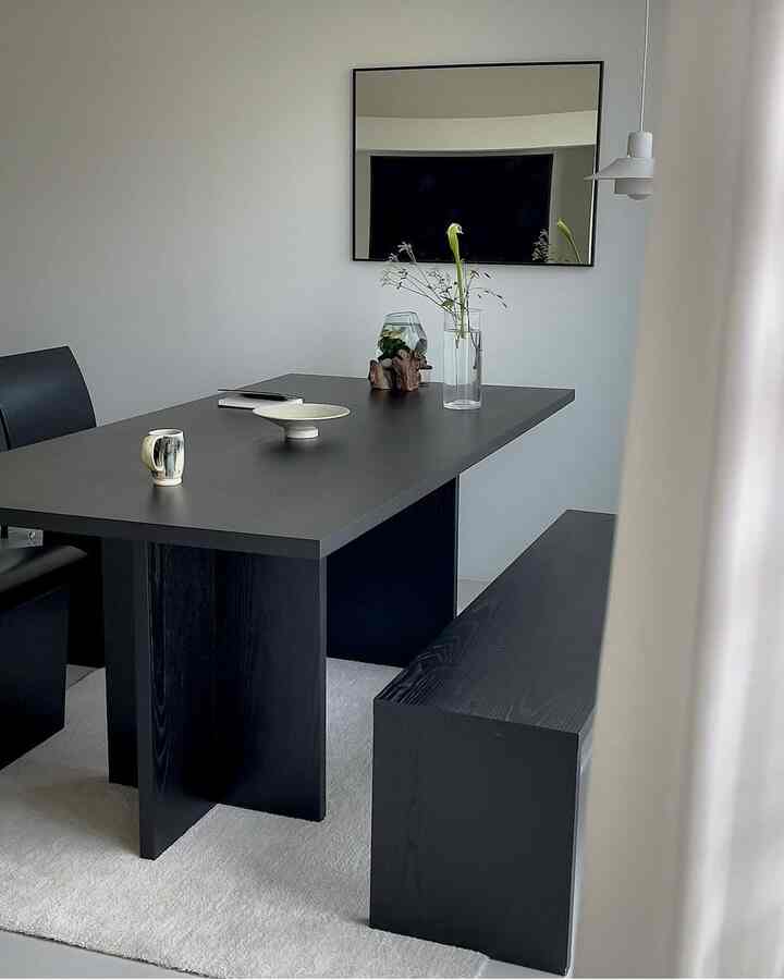 A modern dining room in black and white tones featuring a black dining table, bench, armchair, a large mirror on the wall, and vases with flowers on the table