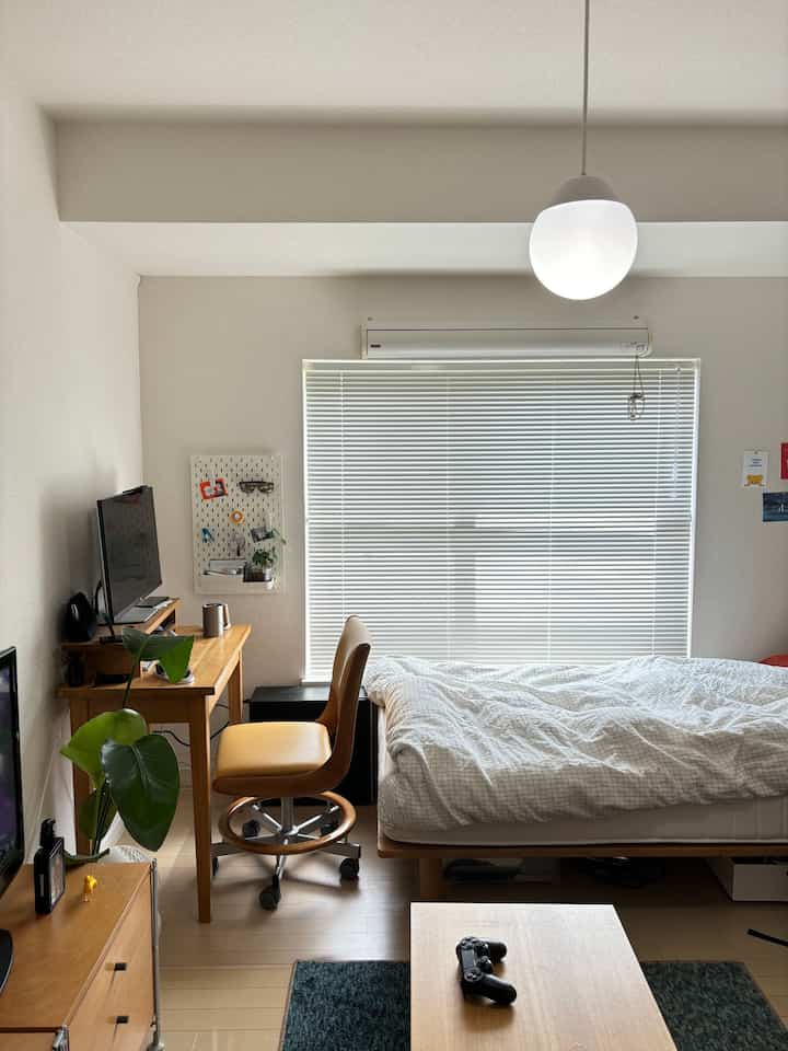 明るい木目調とナチュラルカラーの1K寝室、ペンダントライトと机、植物が配された雰囲気のある空間