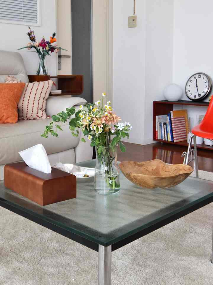 A white and natural toned living room featuring a glass vase with flowers and a wooden bowl on a glass coffee table in a simple setting