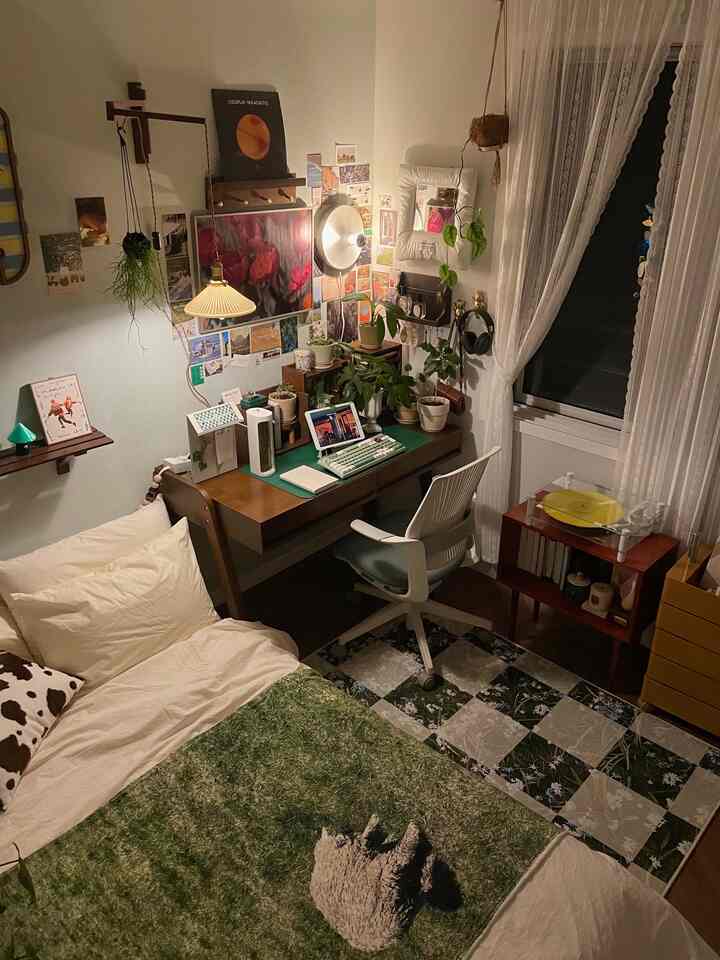 ナチュラルな色調の小さな寝室のホームオフィス、木製の机と緑のラグ、多様な照明と棚が配置された雰囲気のある空間