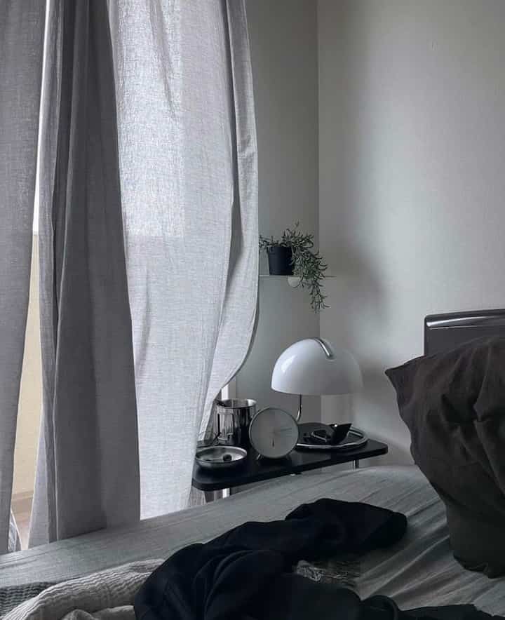 A simple, calm bedroom featuring beige tone curtains and a nightstand with a lamp, creating a cozy atmosphere
