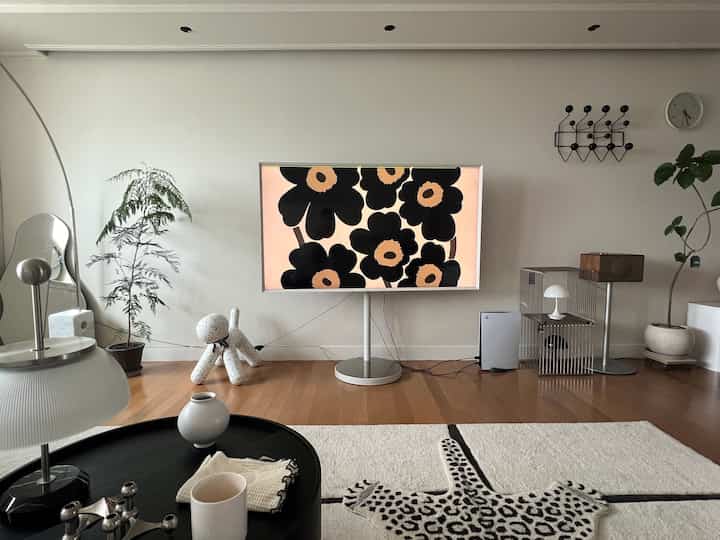 A modern living room in white and brown wood tones, featuring a central TV stand, round table, and plants