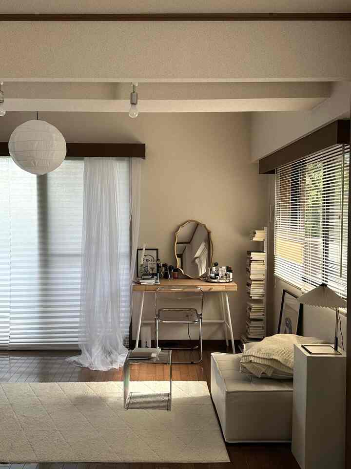 ナチュラルカラーの小さな部屋、中ほどにデスクとクリアチェア、左側にカーテンがあり、シンプルで居心地の良いインテリア