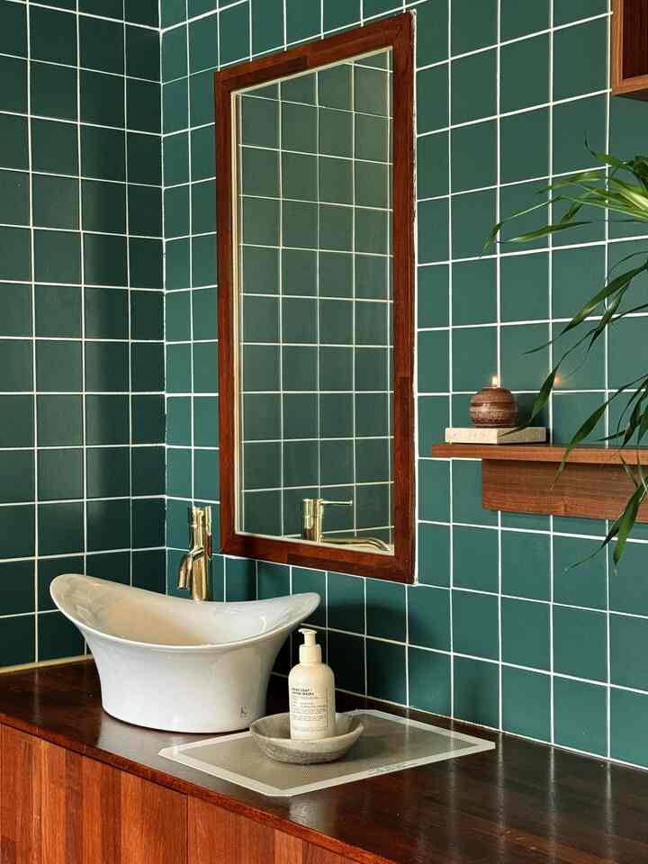 濃い緑のタイルと木目調の鏡、洗面台が配置された落ち着いたバスルーム、小さなキャンドルと植物がアクセントのインテリア