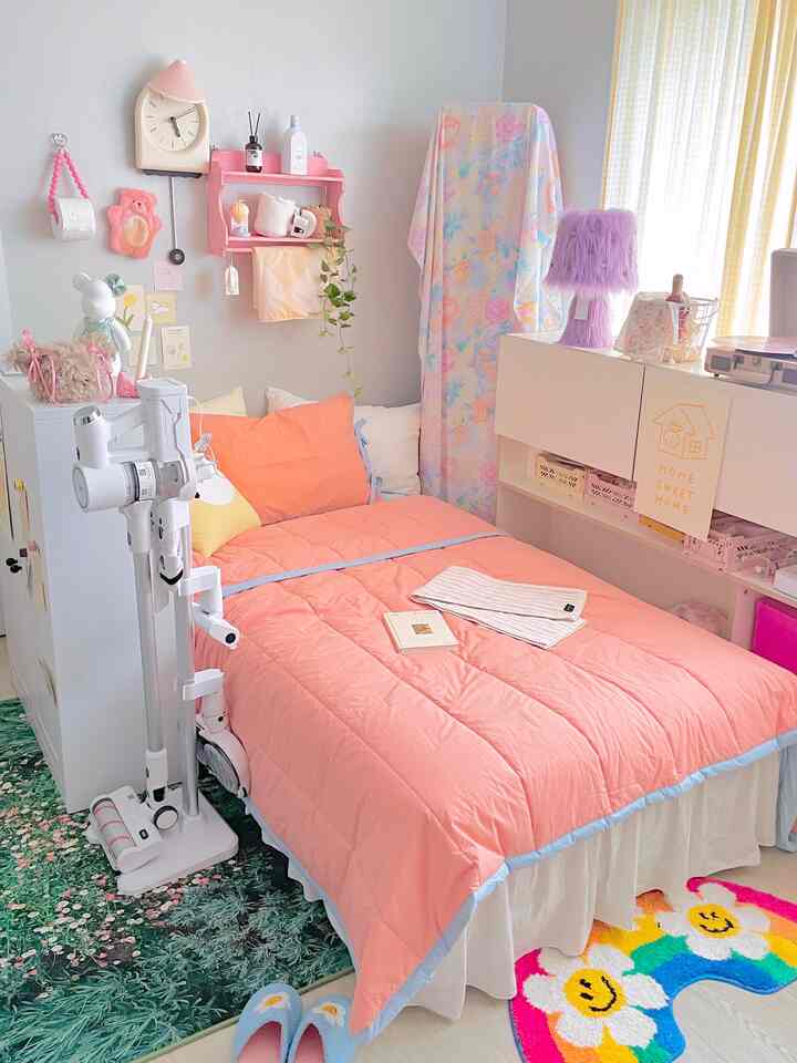 パステル調のコンパクトな寝室、中央にピーチピンクのベッドと白い家具、多彩なラグと紫のテーブルランプが配置された印象的な空間