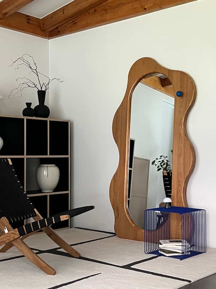 Natural wood tone living room with white walls, featuring a wavy wooden mirror and a modern lounge chair