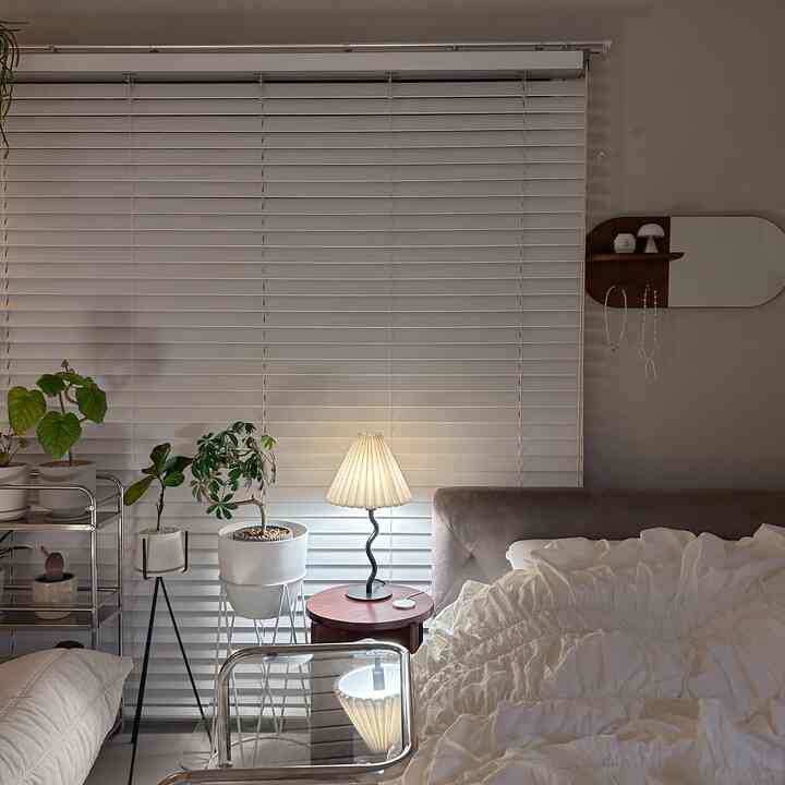 Cozy bedroom in soft white and brown tones featuring nightstand, table lamp, and multiple plants