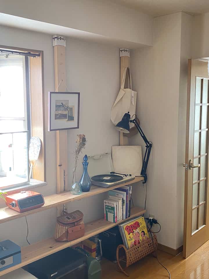 Natural-toned compact single household room with white walls, brown wooden floor, shelving holding books, record player, and desk lamp