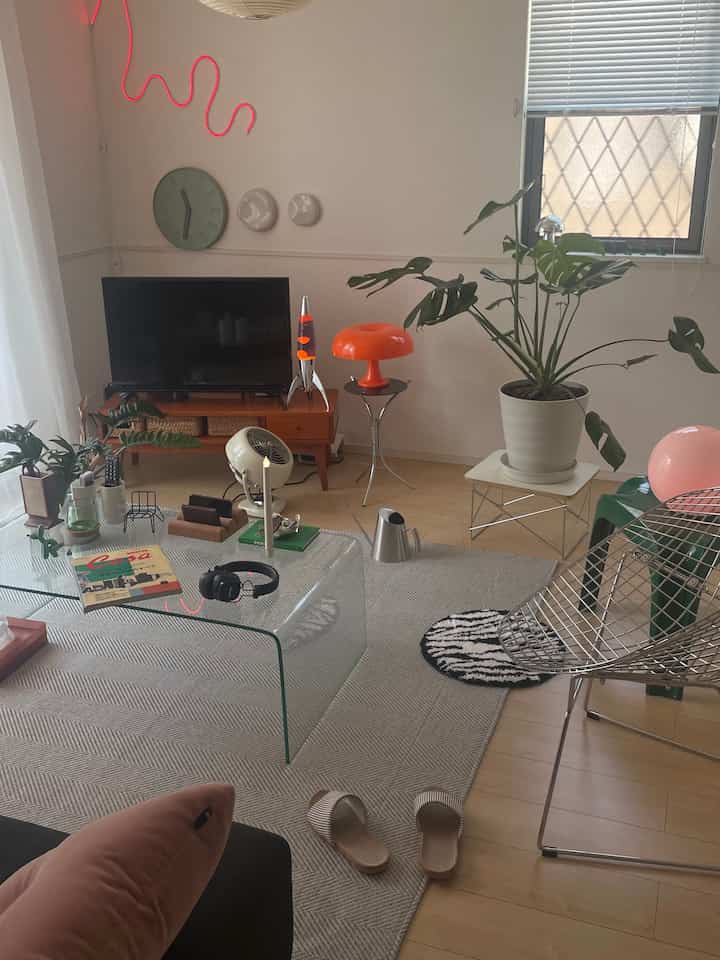 A bright wood-floor living room with white walls, featuring a transparent glass coffee table, an orange table lamp, and multiple green plants in a cozy mid-century modern style.