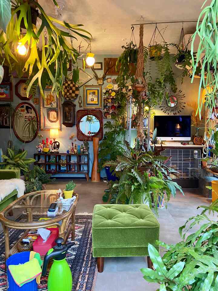 緑を基調としたリビング、豊かな観葉植物とミッドセンチュリーモダンスタイルのオットマン、ヴィンテージの壁飾りが配置された雰囲気のあるインテリア