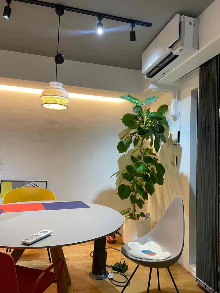 ナチュラルカラーのコンパクトなリビング、丸テーブルとカラフルな椅子、大型の植物とエアコンが配置されたモダンなインテリア