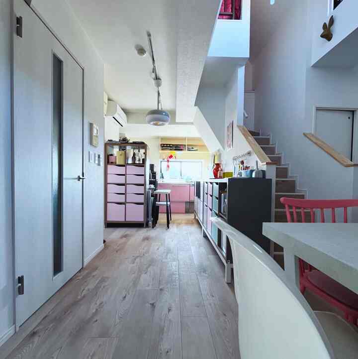 Bright white walls and light brown wood flooring define a compact two-person household space featuring stairs, storage shelves, and a cozy living-dining-kitchen area.