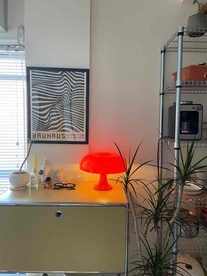 明るい白い壁とミッドセンチュリーモダンのポスターがあるリビング、丸い赤いテーブルランプと緑の植物が配置されたシンプルな空間