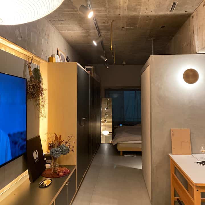 モダンなスタイルの狭い寝室、黒いワードローブと木目調のコンソールテーブル、コンクリート天井と壁照明が配置された落ち着いたインテリア