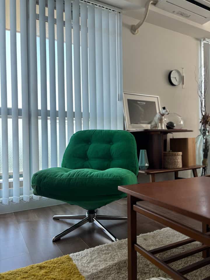 明るいブルーのブラインドと木目調家具が配置されたミッドセンチュリーモダンのリビング、緑のアームチェアと黄色のラグが特徴的な空間