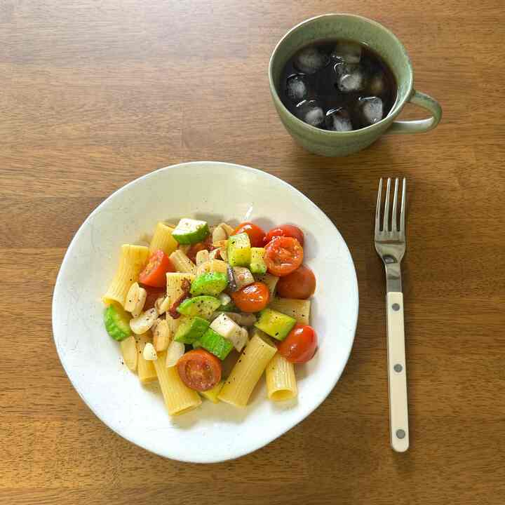 温かみのあるウッドトーンのダイニングテーブルに、白いパスタボウルと野菜のパスタ、隣にアイスコーヒーとフォークが置かれた、雰囲気のあるおうちカフェ空間