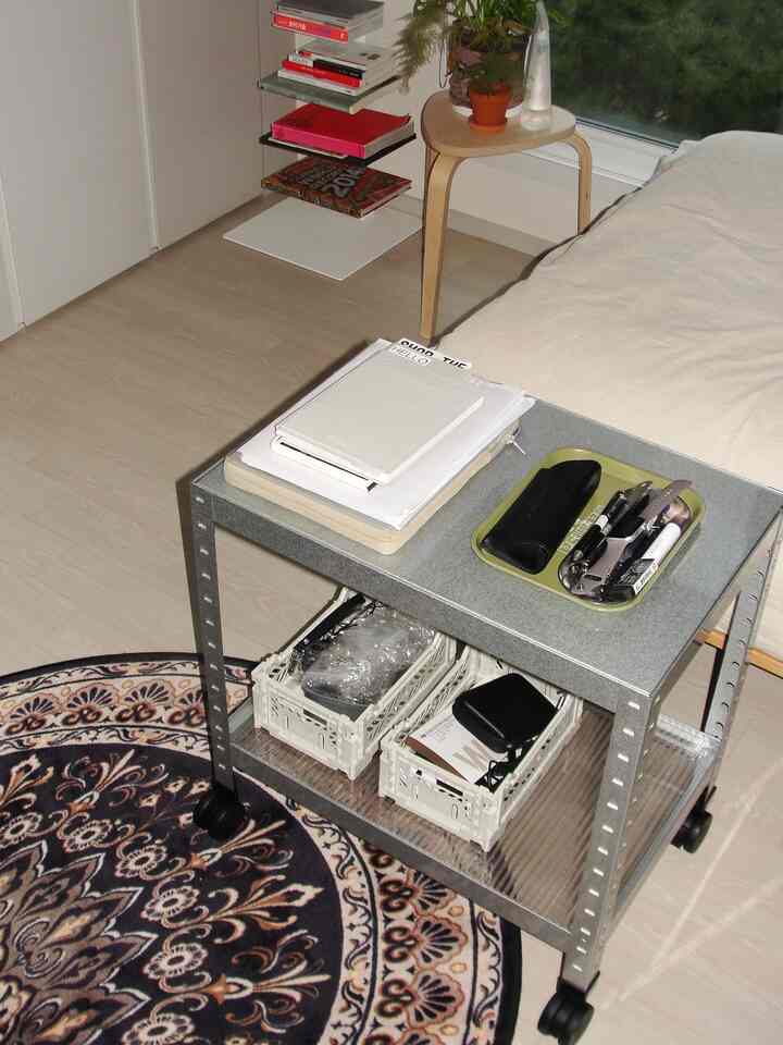 ナチュラルな色調の小さな寝室、クリーム色の寝具と移動式の金属製収納カートが配置された雰囲気のあるインテリア