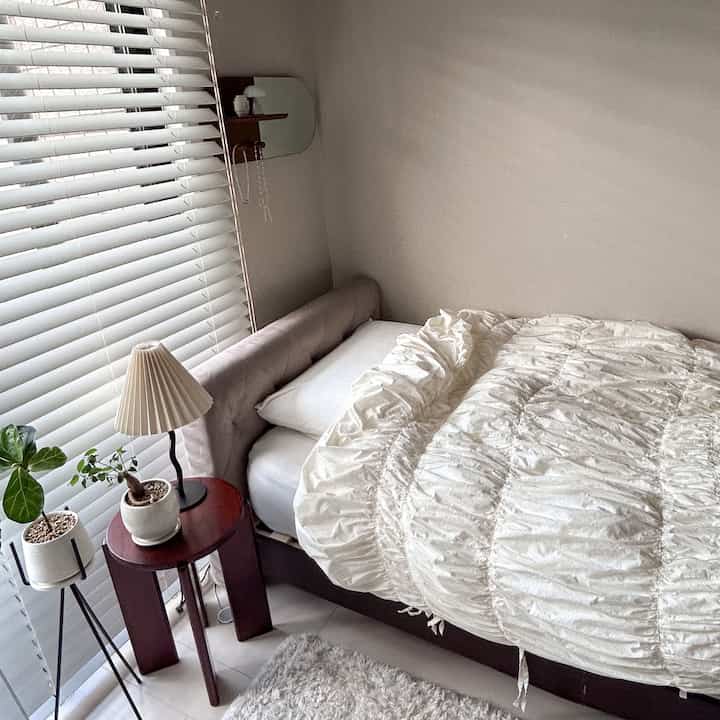 ベージュとホワイトのトーンの小さな寝室、ベルベットヘッドボードベッドと丸いナイトテーブル、植物が配置された温かみのある自然なインテリア