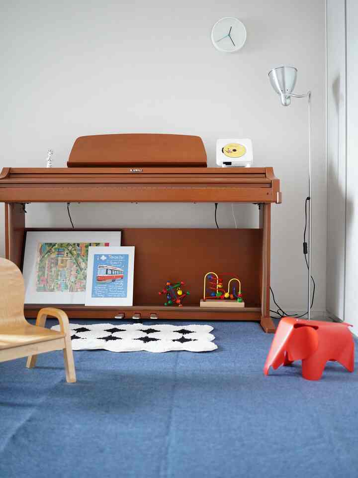 茶色のピアノと青いカーペットの子供部屋、カラフルな玩具と赤い象のスツールが置かれた印象的な空間
