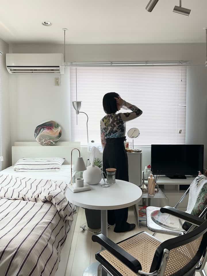 White-toned 1K bedroom space featuring striped bedding, round table, and horizontal blinds with a simple interior