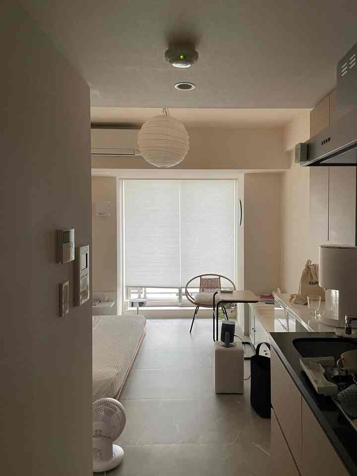 明るいベージュと白の色調のワンルーム、自然素材の家具とシンプルなキッチンが配置された落ち着いた空間
