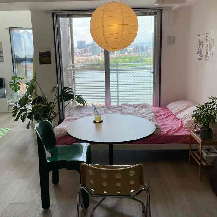 ピンクの寝具と丸いテーブル、木目調の床と観葉植物が配された明るいワンルームのインテリア