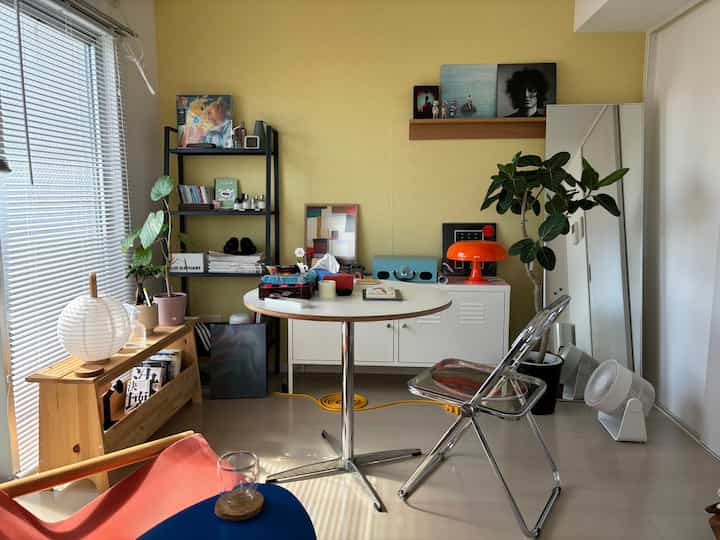 明るい黄色の壁と白い家具が調和する7畳のワンルーム、透明なスツールや植物、オレンジのテーブルランプがアクセントの温かみのある空間