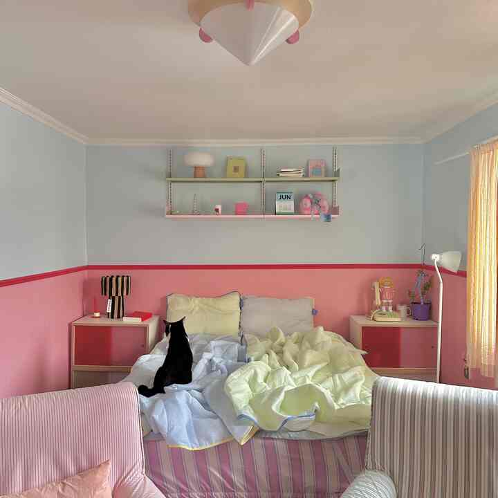 ピンクと白のトーンの小さな寝室、ベッドに黒猫が座り、両側にナイトテーブルと可愛らしい小物が配置された温かな空間