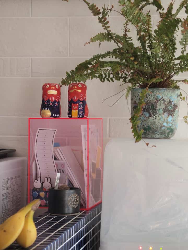 白い壁と黒いタイルのキッチン調理スペース、透明な収納ボックスに赤い置物と緑の植物が配置された落ち着いたインテリア