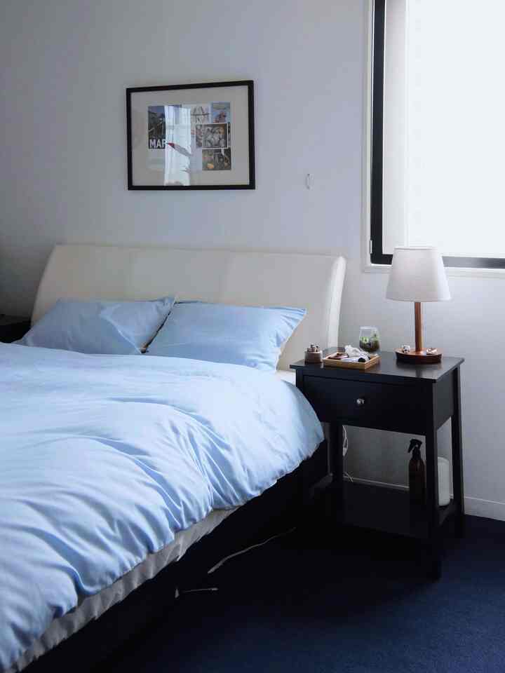 白と青の色調が調和した寝室、右側のナイトテーブルにテーブルランプと青い寝具が配され、落ち着いた雰囲気のインテリア