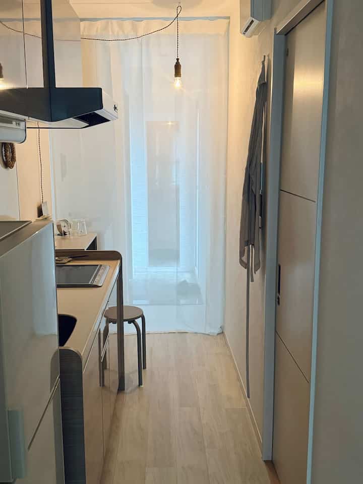 Bright white and natural-toned long 1K kitchen space featuring a simple stool and sheer curtain, creating a minimalistic atmosphere