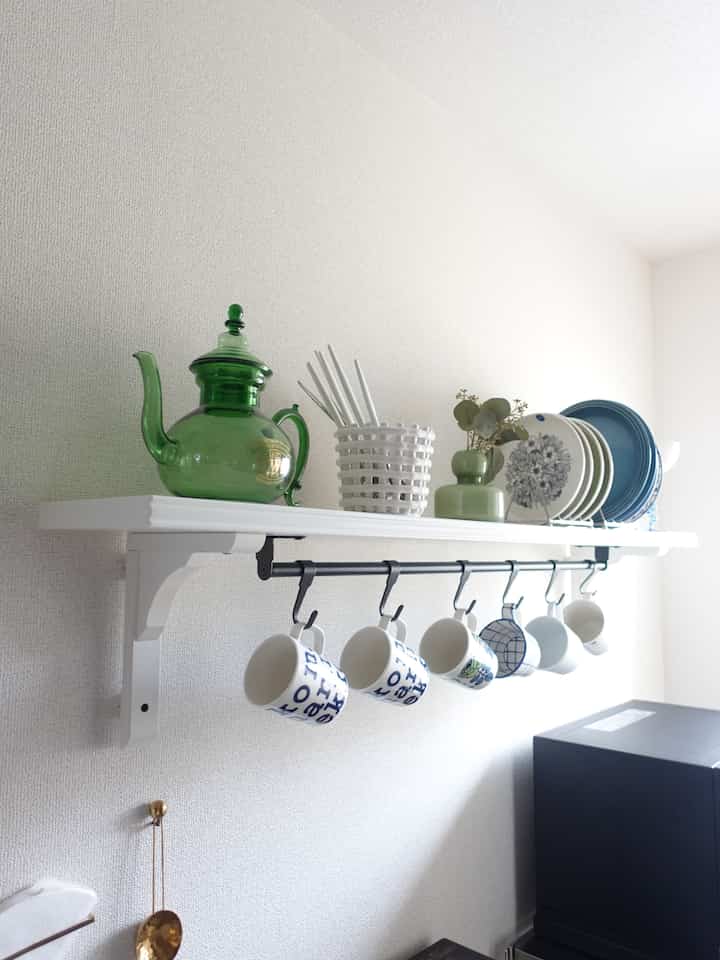 白い壁と北欧風の壁棚にマグカップや皿、緑のガラス製のポットが並ぶ清潔感のあるキッチン