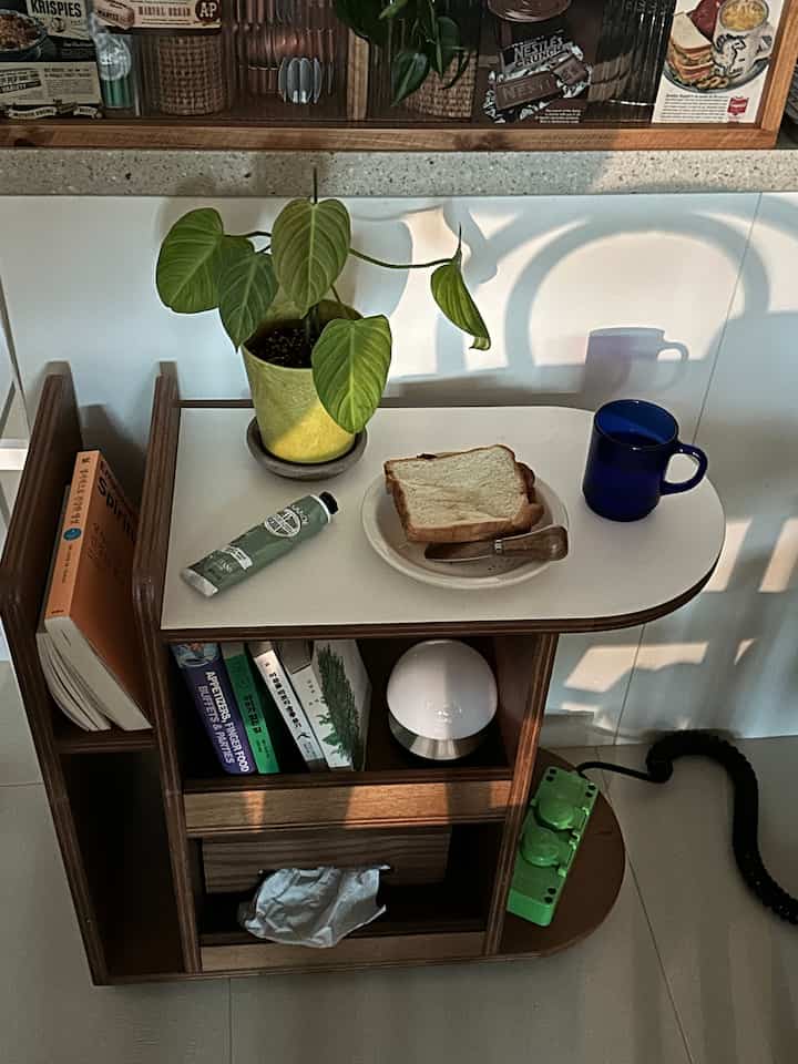 ナチュラルトーンの木目調トロリーが置かれたおうちカフェ風の小さな空間、植物とパンとカップが配置されている