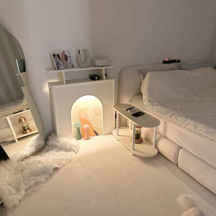 白とベージュのトーンが調和した二人暮らしの寝室、ファブリックベッドと暖炉コンソール、ラグが配されたモダンで落ち着いたインテリア